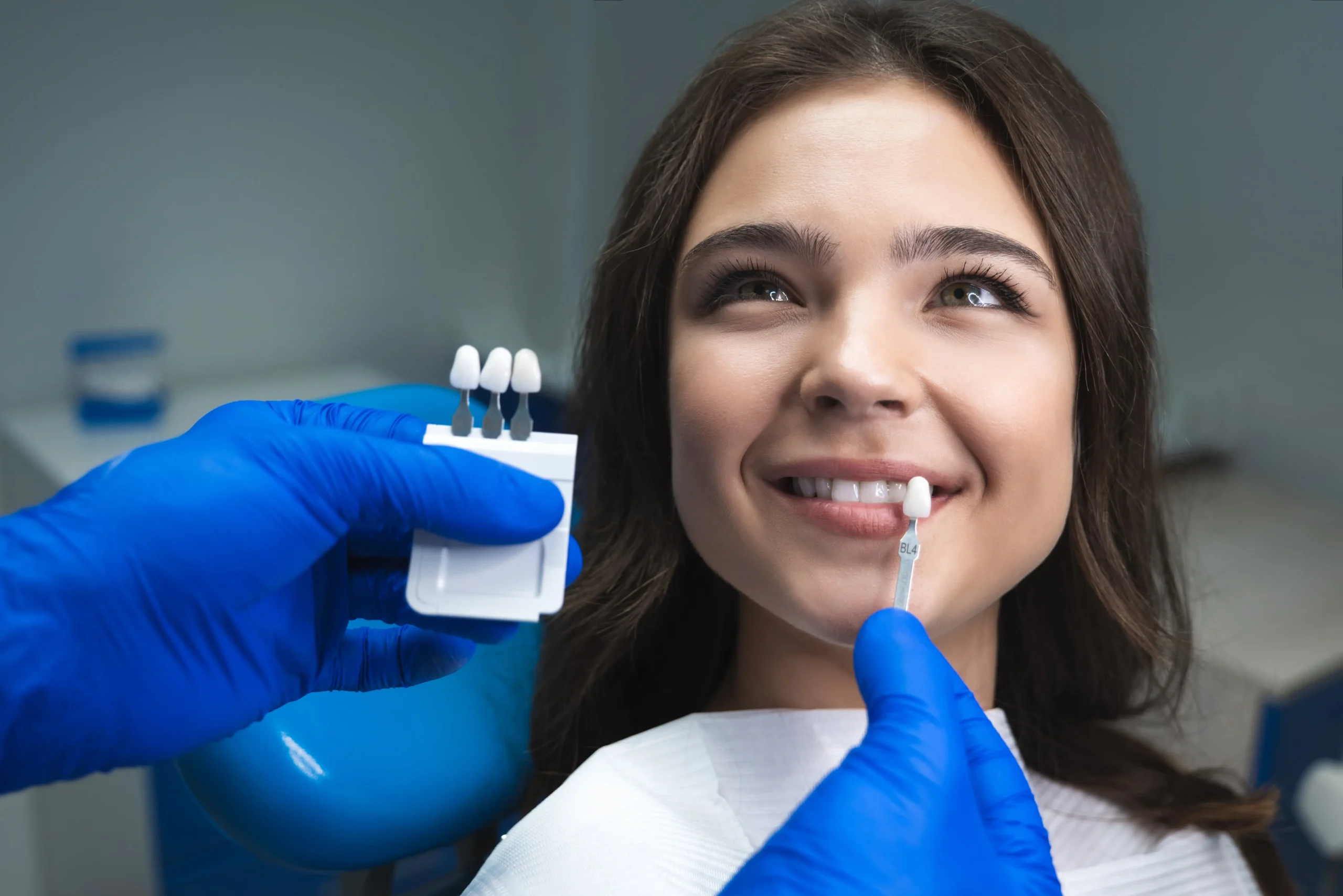 A woman at the dentist. The dentist is checking the woman's teeth color using a teeth shading palette.
